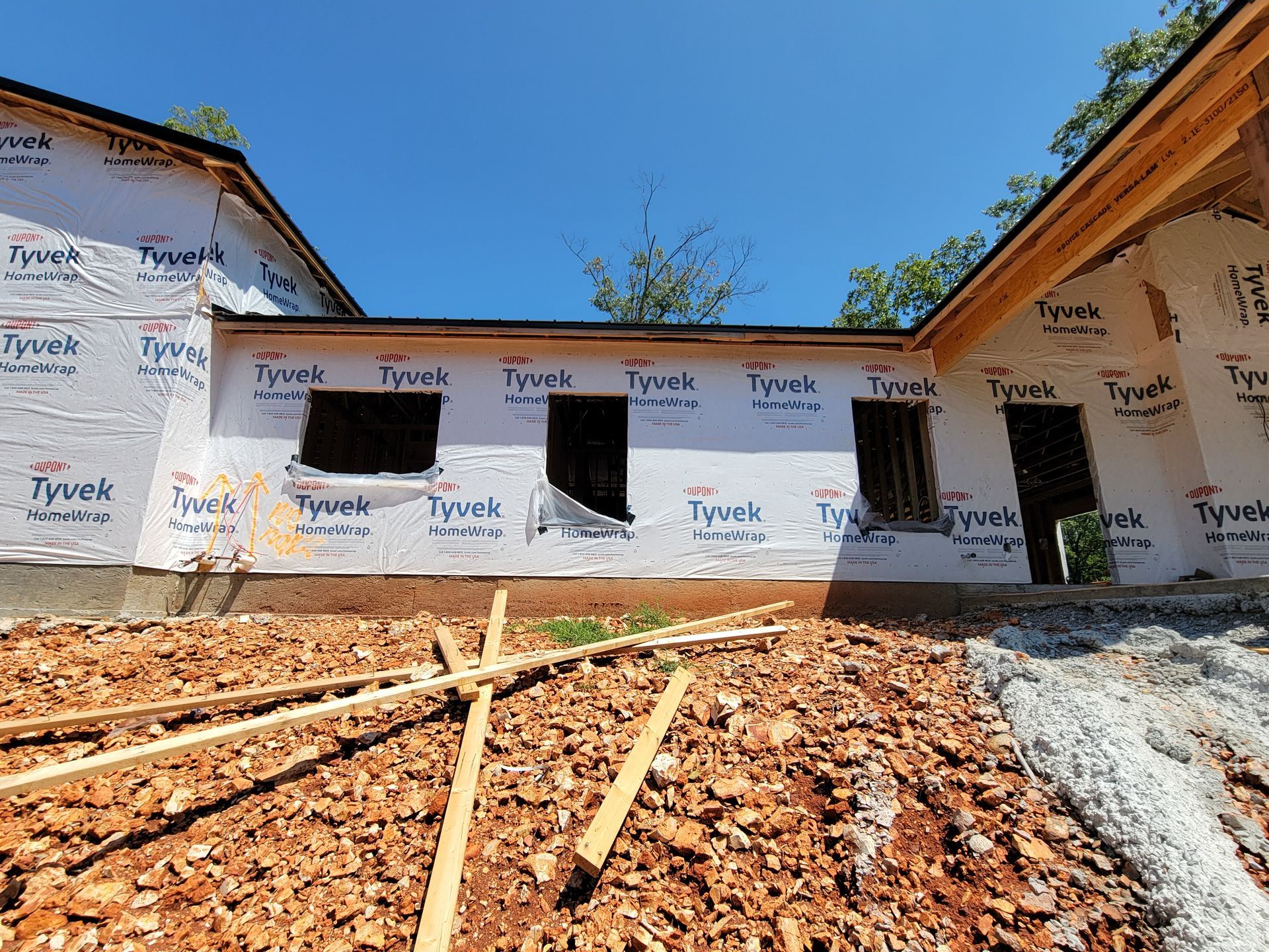 Partially built house wrapped in Tyvek, with unfinished dirt yard under a blue sky