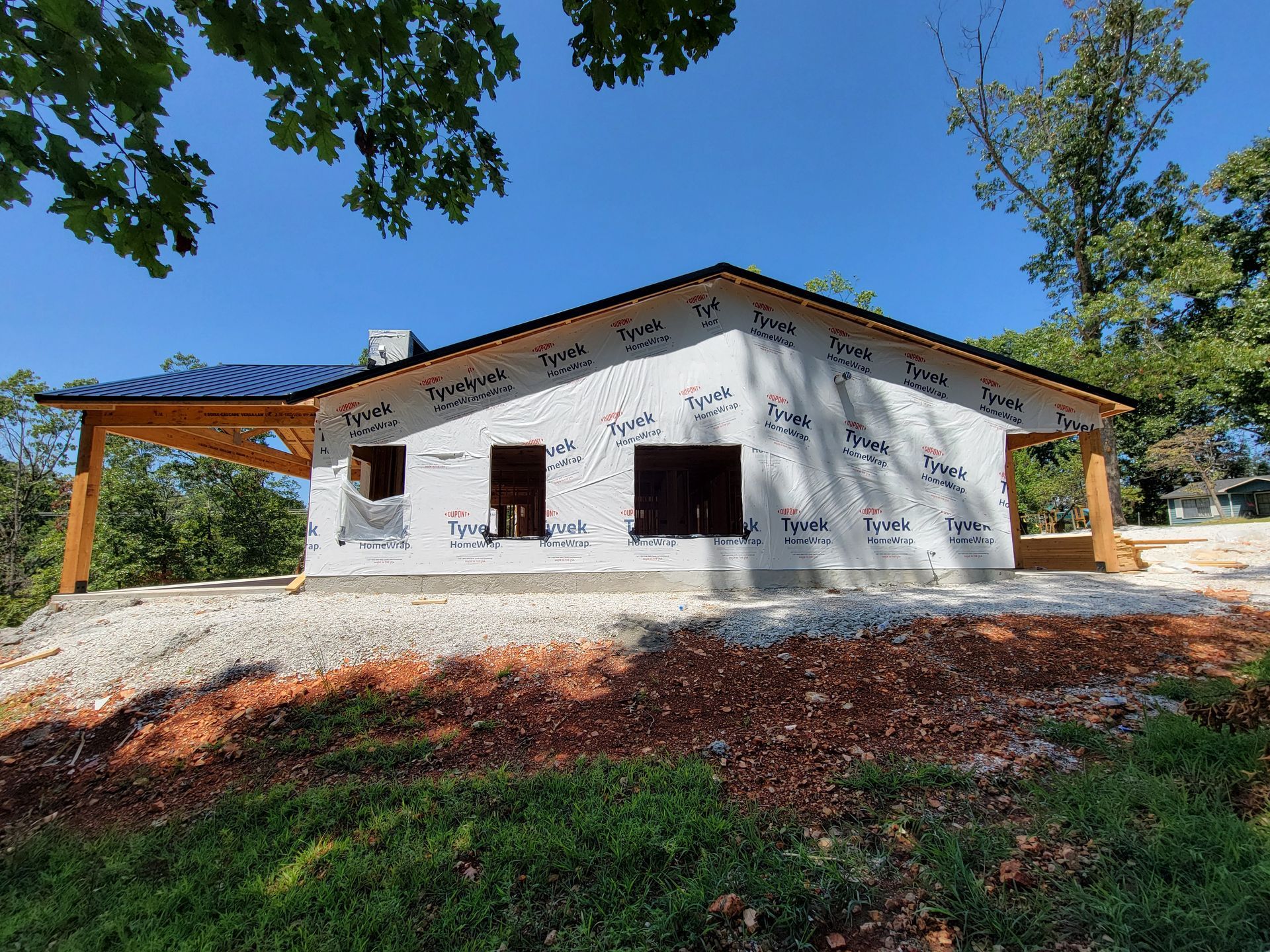 White house with brown trim on a wooded hillside under a blue sky, framed by trees and green grass