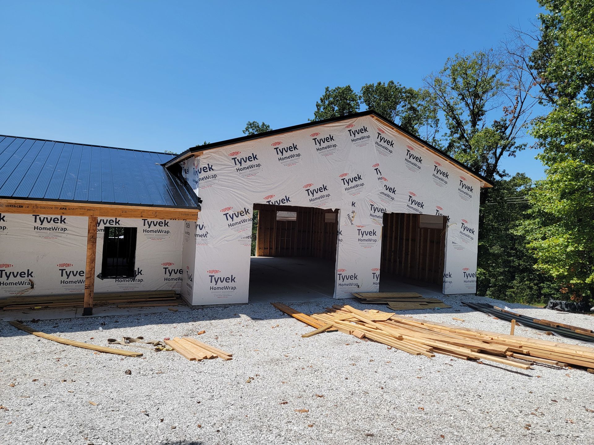 Partially built white house with open garage bays, blue roof, and gravel driveway under a clear sky