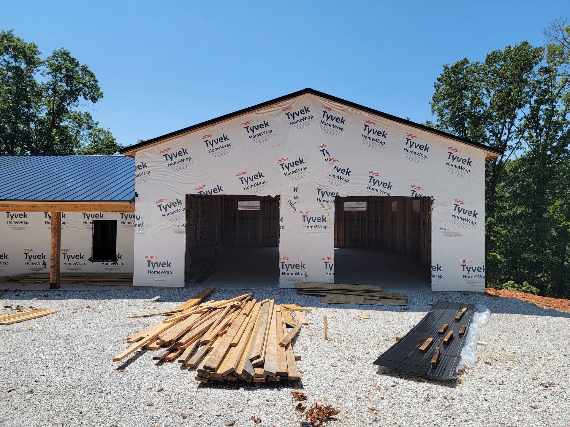 Partially built garage with blue metal roof, wrapped siding, and lumber stacked on gravel in front