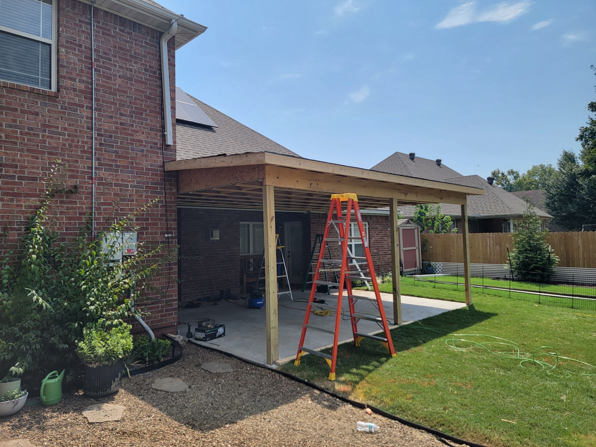 Backyard patio roof under construction with a red ladder beside a brick house