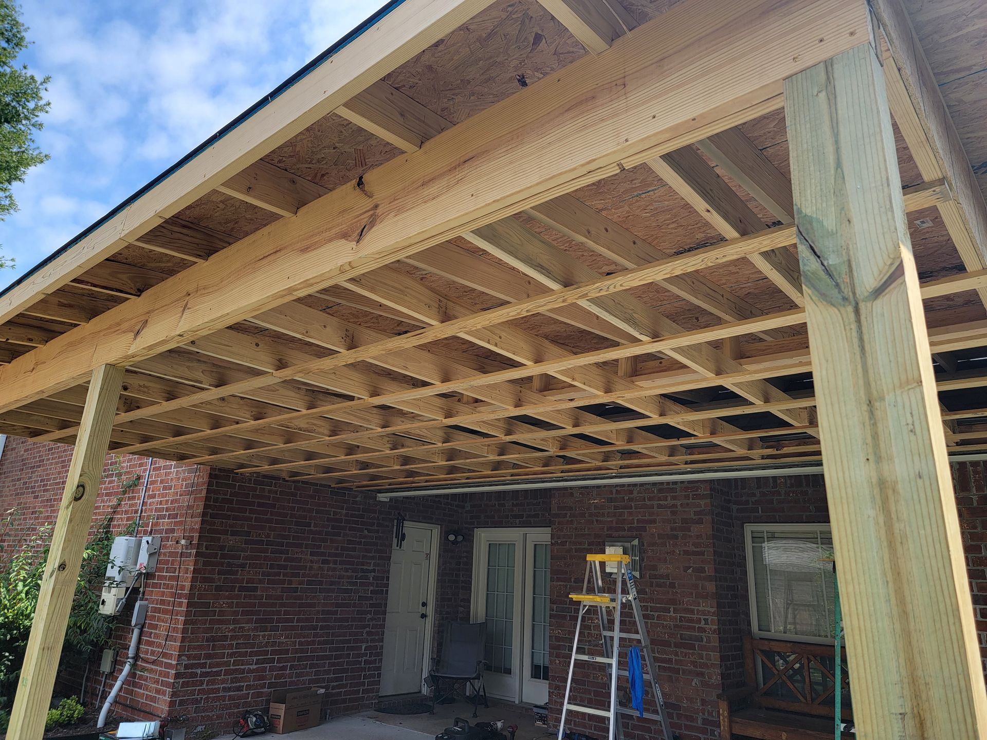 Partially built wooden porch roof with exposed framing and a ladder under a brick house