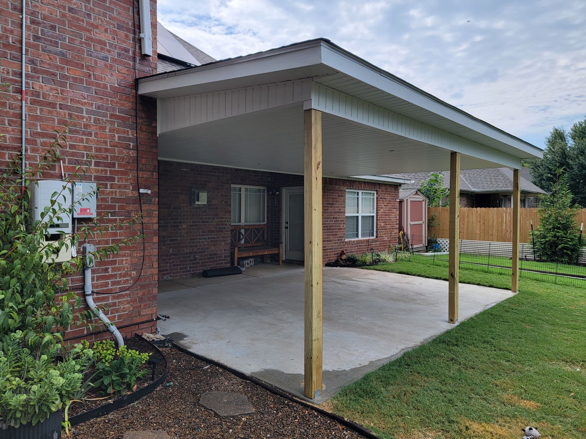 Covered concrete patio beside a brick house, with a white roof and wooden support posts.
