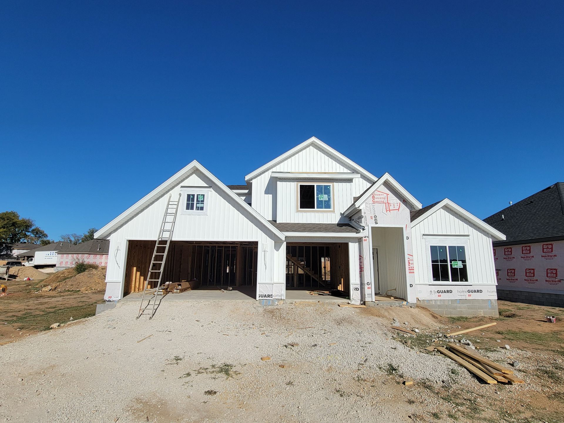 New white house under construction with open garage, ladders, and gravel driveway under a clear blue sky