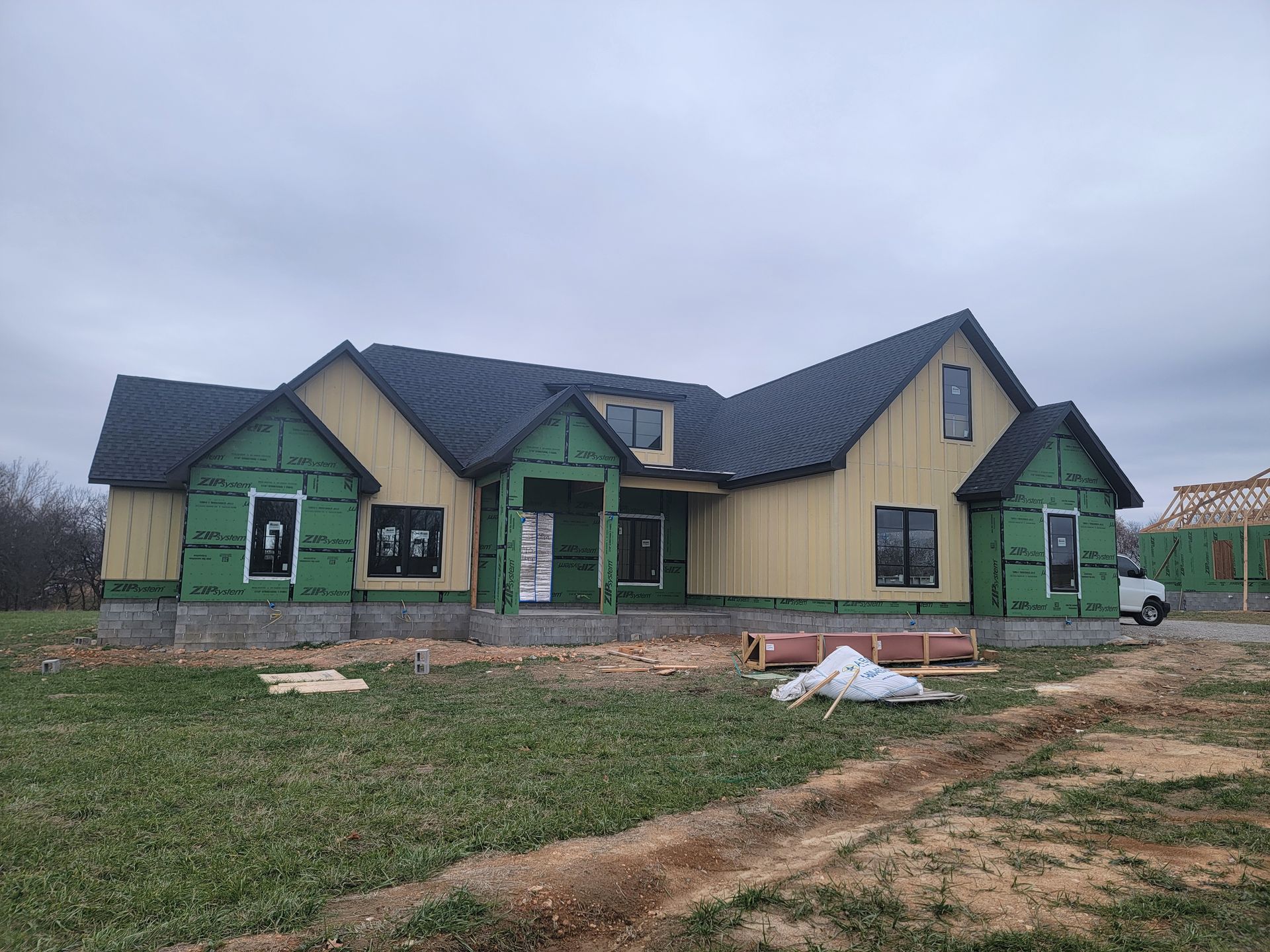 Partially built green house with dark roof on a muddy lot under an overcast sky