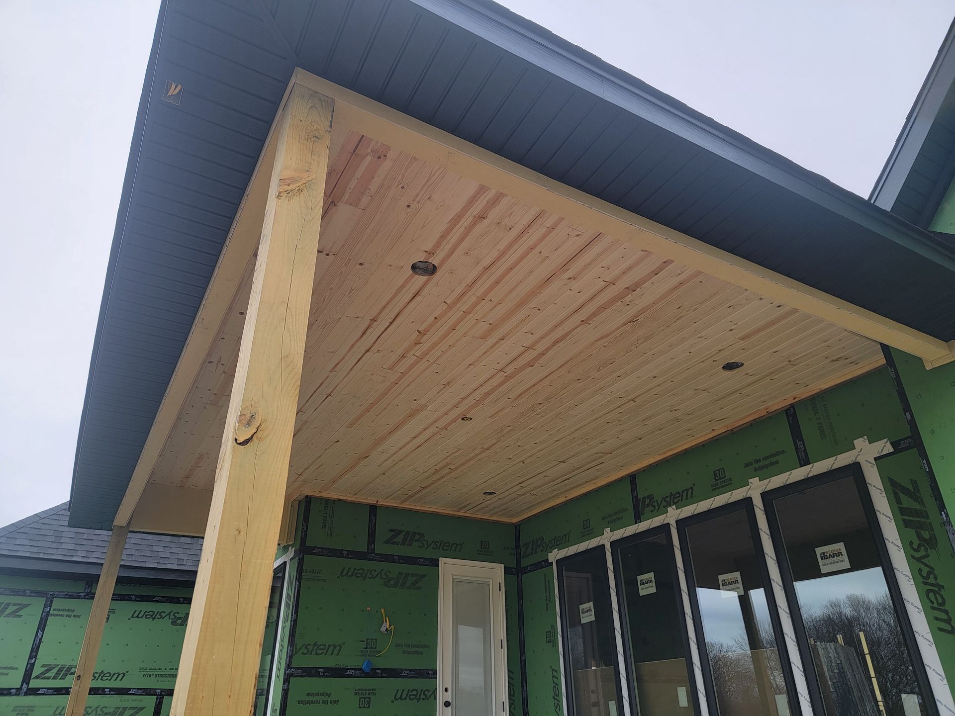 Covered porch with wood ceiling and support post on a green house, showing a glass front door.