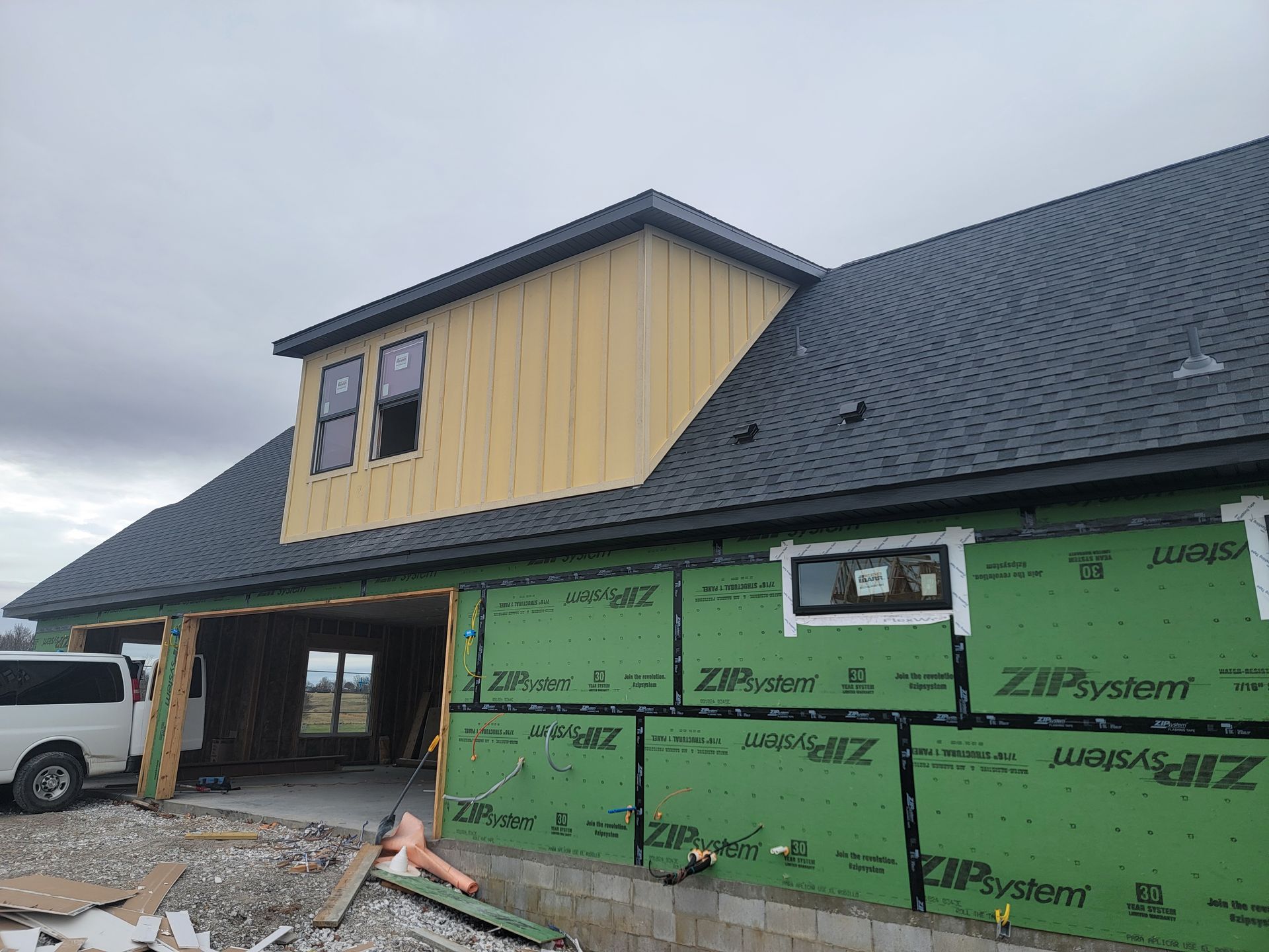 Partly built house with green house wrap, dark roof, and a white van on the left under cloudy skies