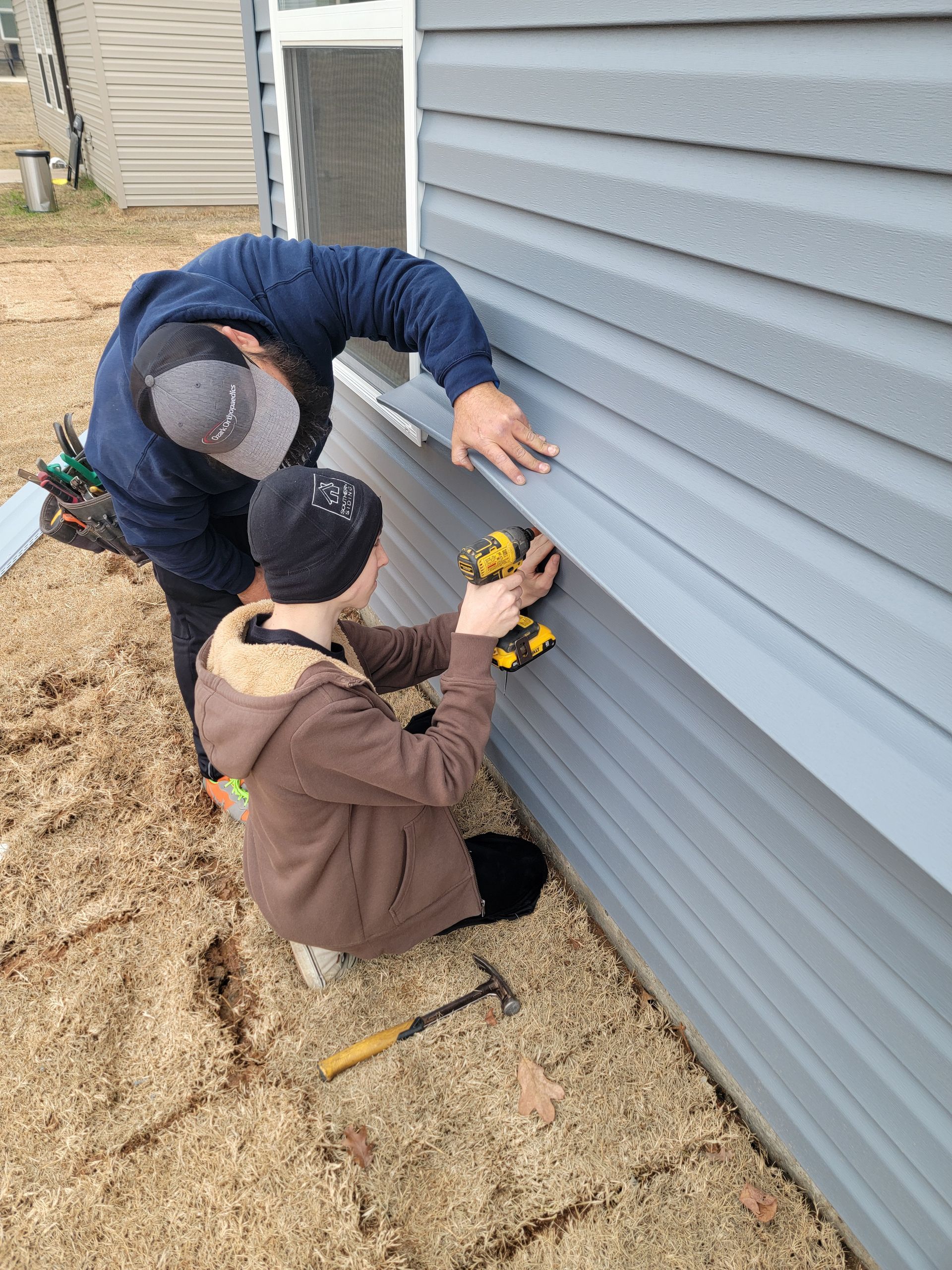 Two people install siding on a house exterior, one kneeling with a drill and one measuring above.