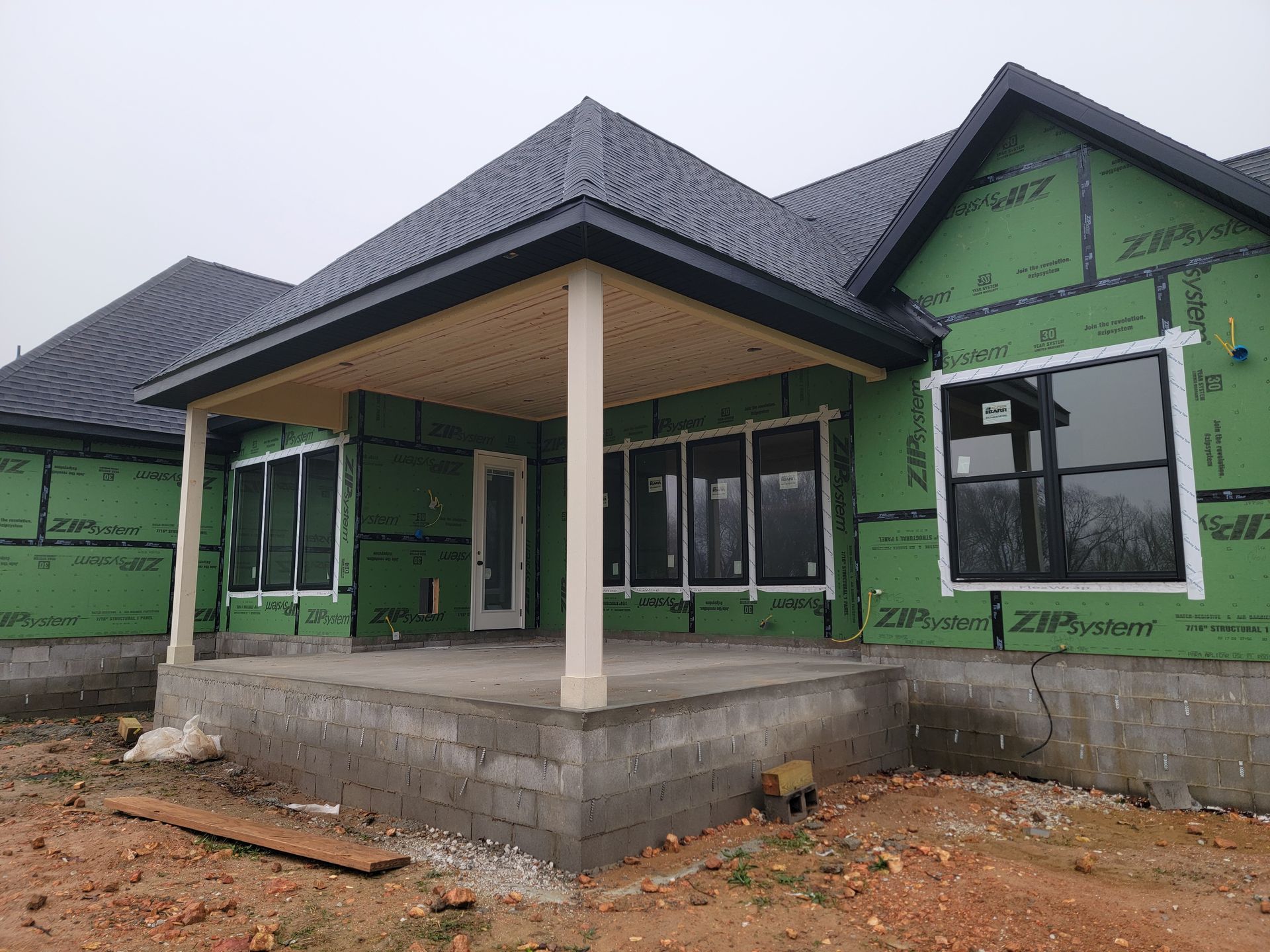 Unfinished house with green wrap, dark roof, and a small front porch on a dirt lot.