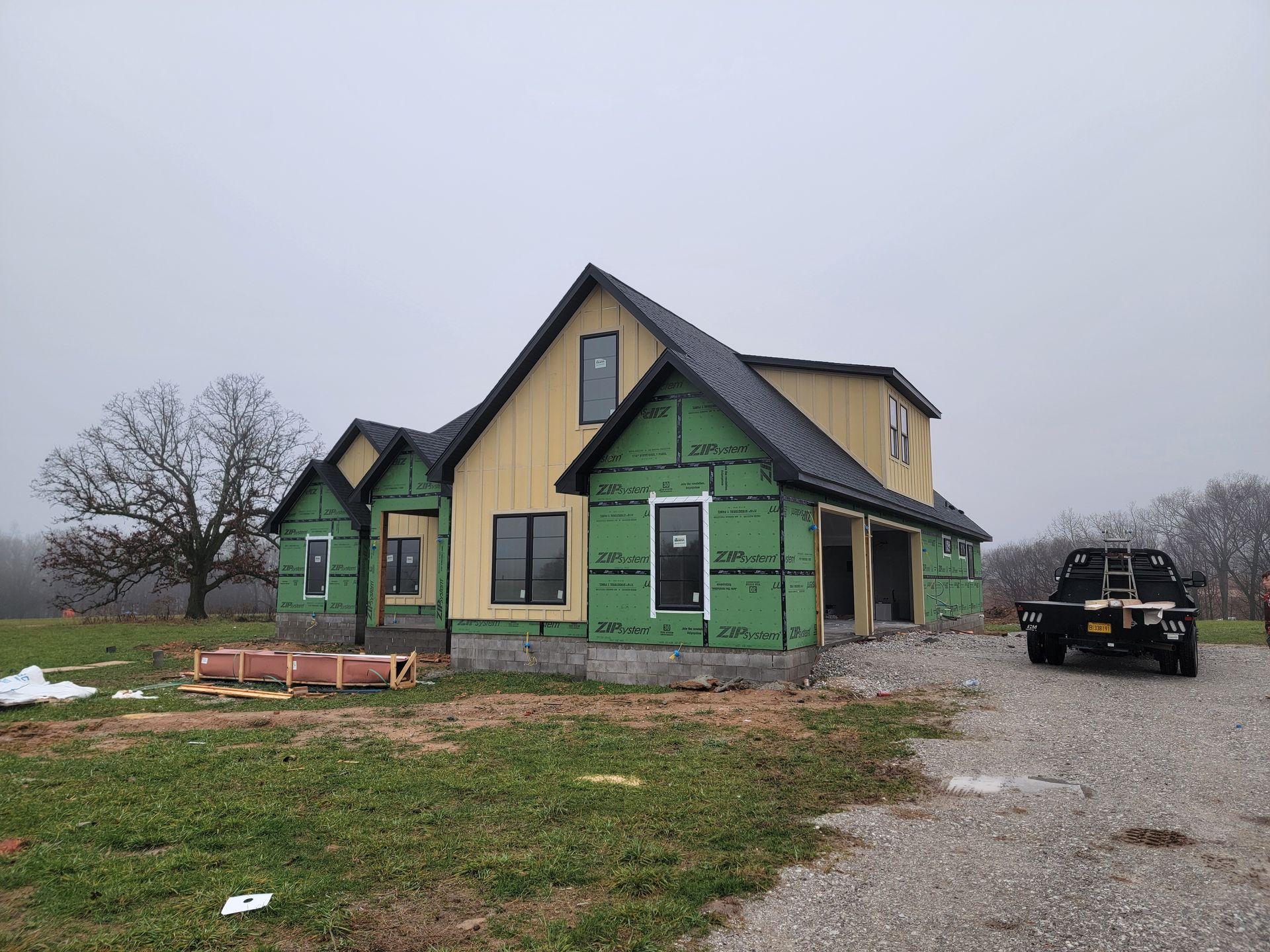 Partially built house with green house wrap, gravel driveway, and black SUV under gray sky