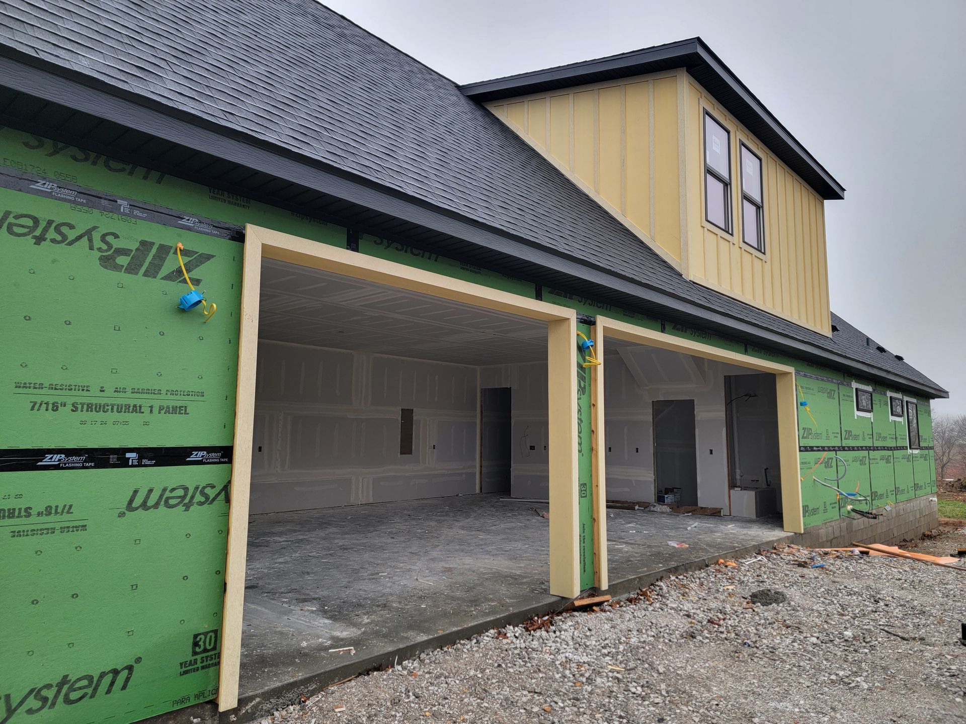 Partially built house with two open garage bays and green exterior wrap under a dark shingled roof
