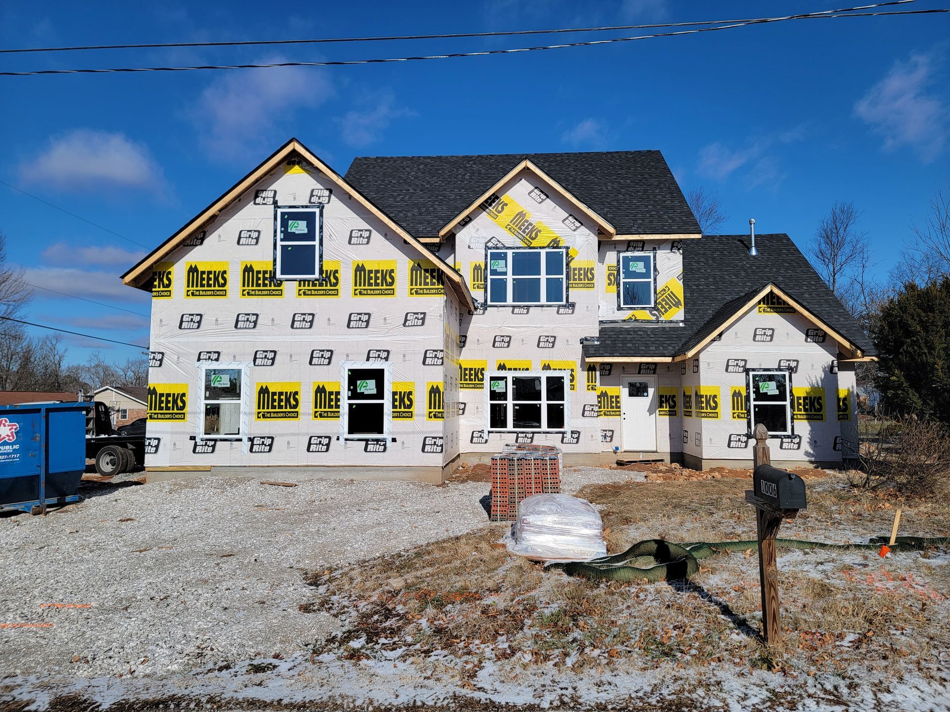 Partially built house with white siding, yellow insulation wrap, and gravel driveway under a blue sky