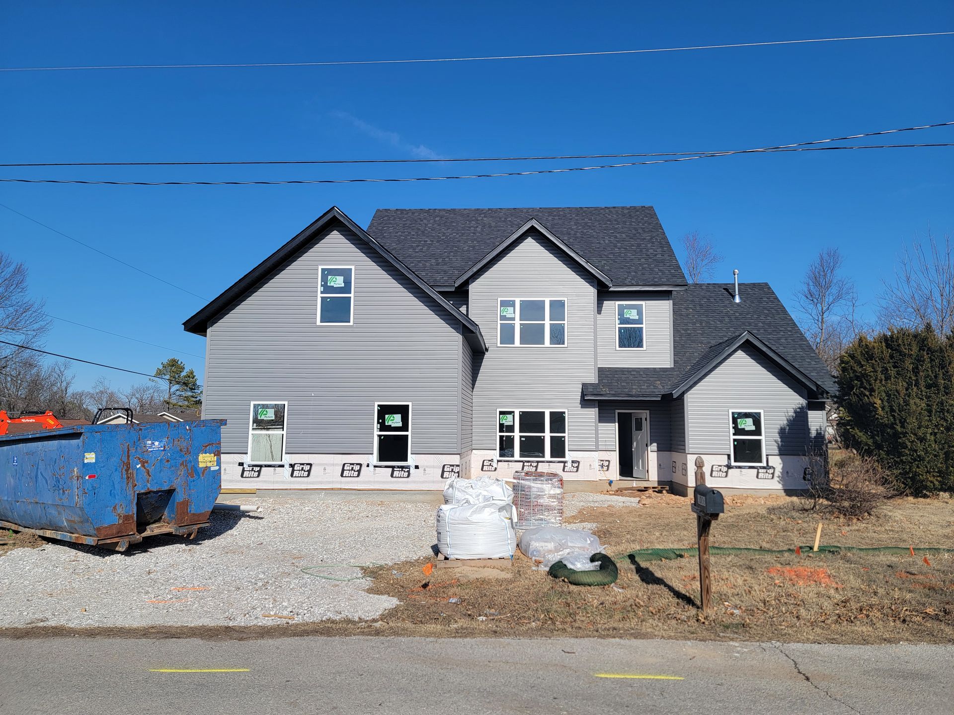 Gray two-story house with a blue dumpster and trash bags in the front yard under a clear sky