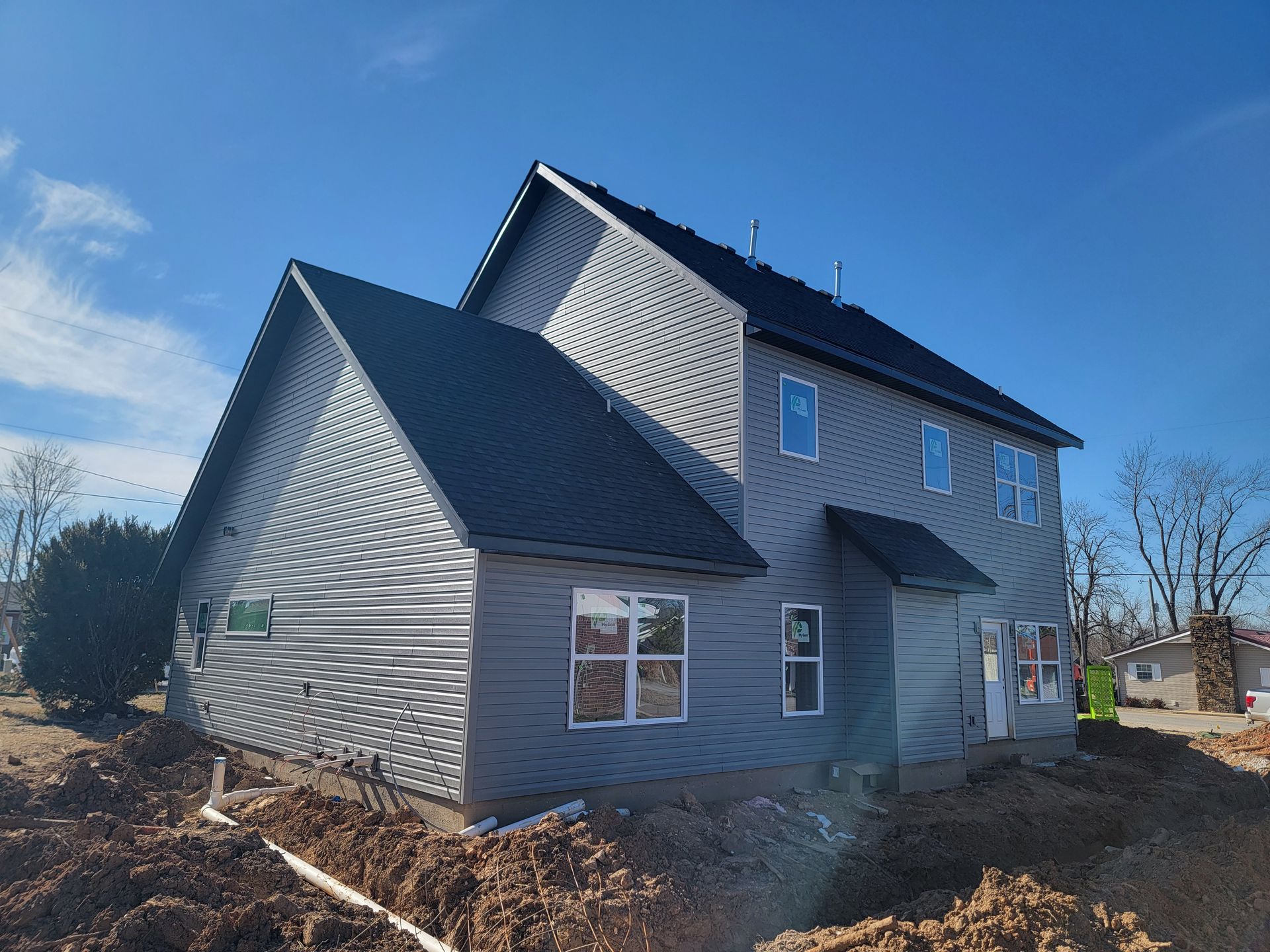 Gray two-story house with dark roof under construction on a dirt lot under a blue sky