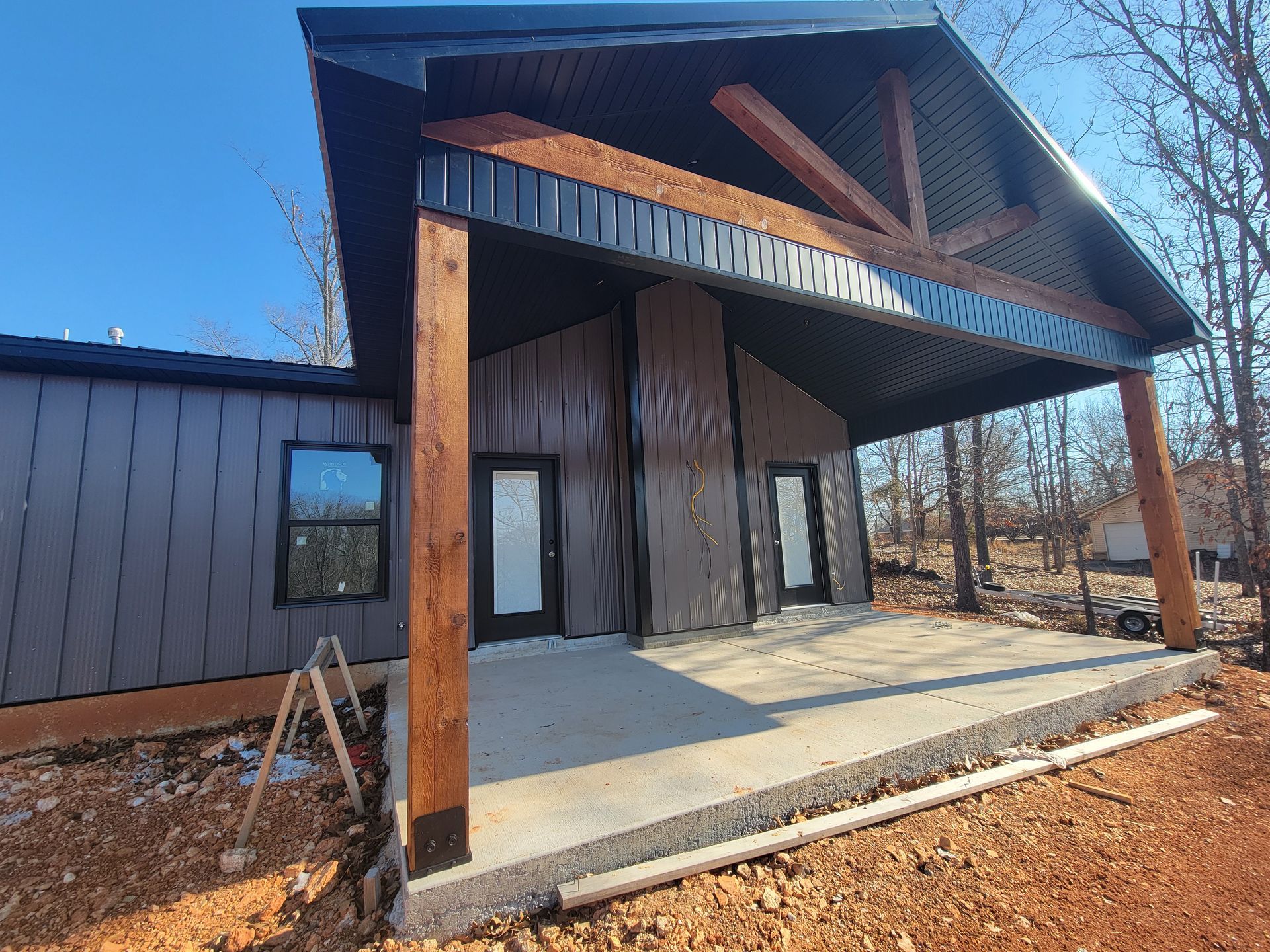 Modern cabin-style house entrance with wood beams, dark siding, and a concrete porch in a wooded area