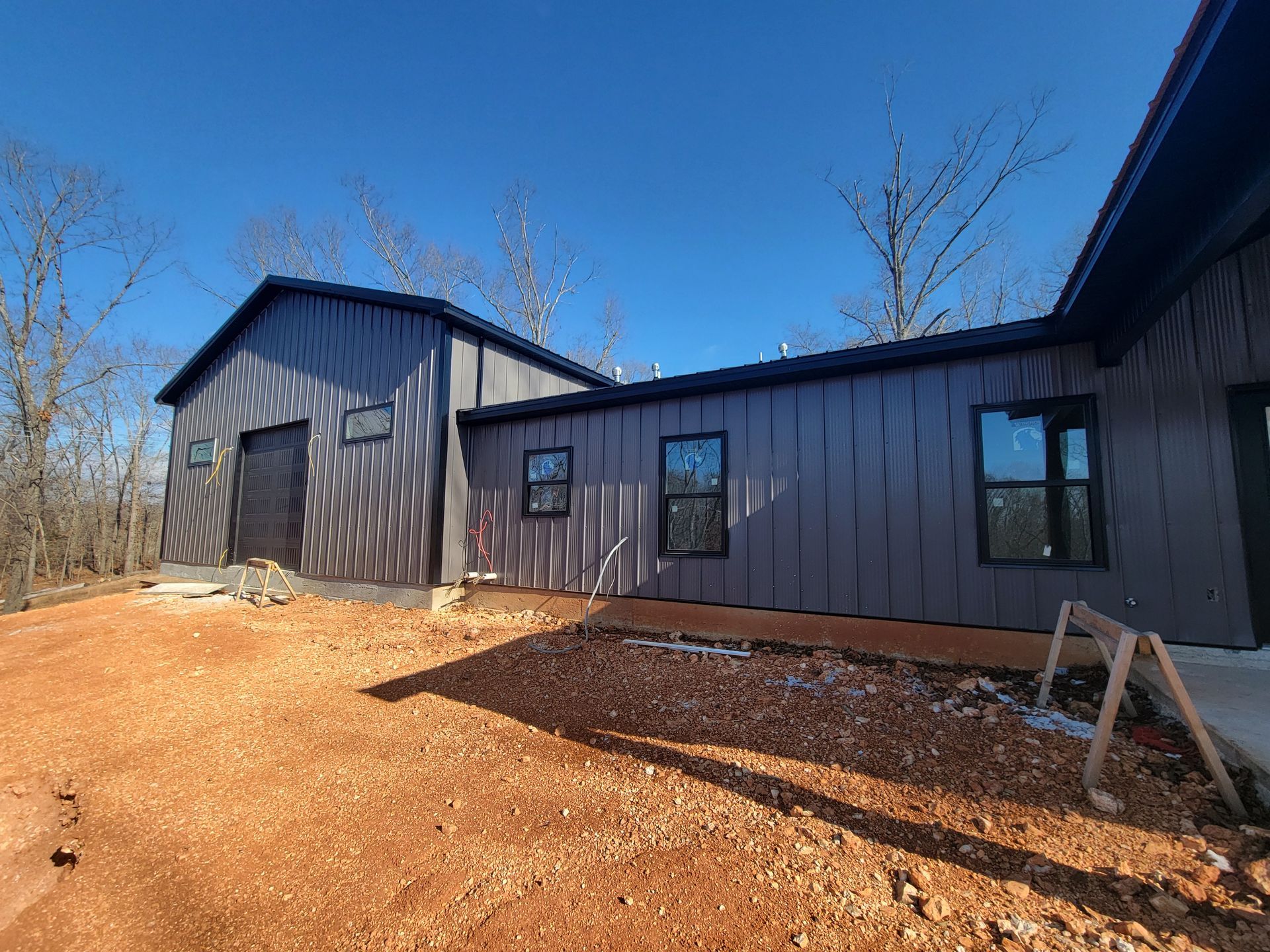 Dark gray house exterior with black windows and a gabled roof on a bare yard under a clear blue sky