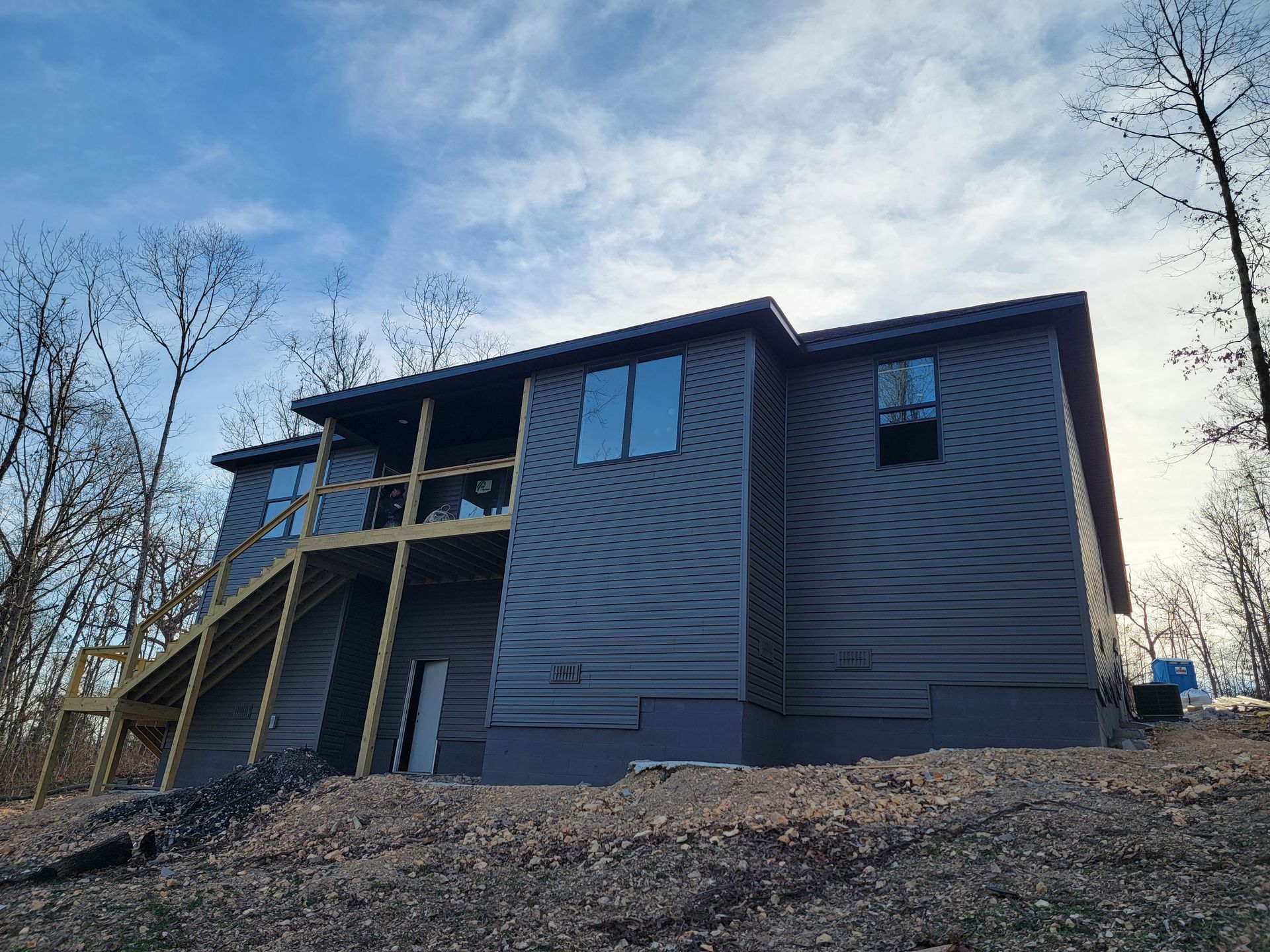 Dark gray two-story house with a deck, viewed from below against a cloudy sky.