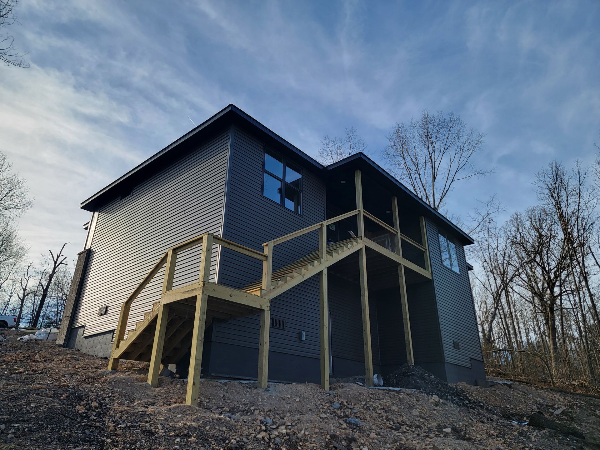 Dark gray house with a wooden ramp and stairs on a wooded hillside under a blue sky