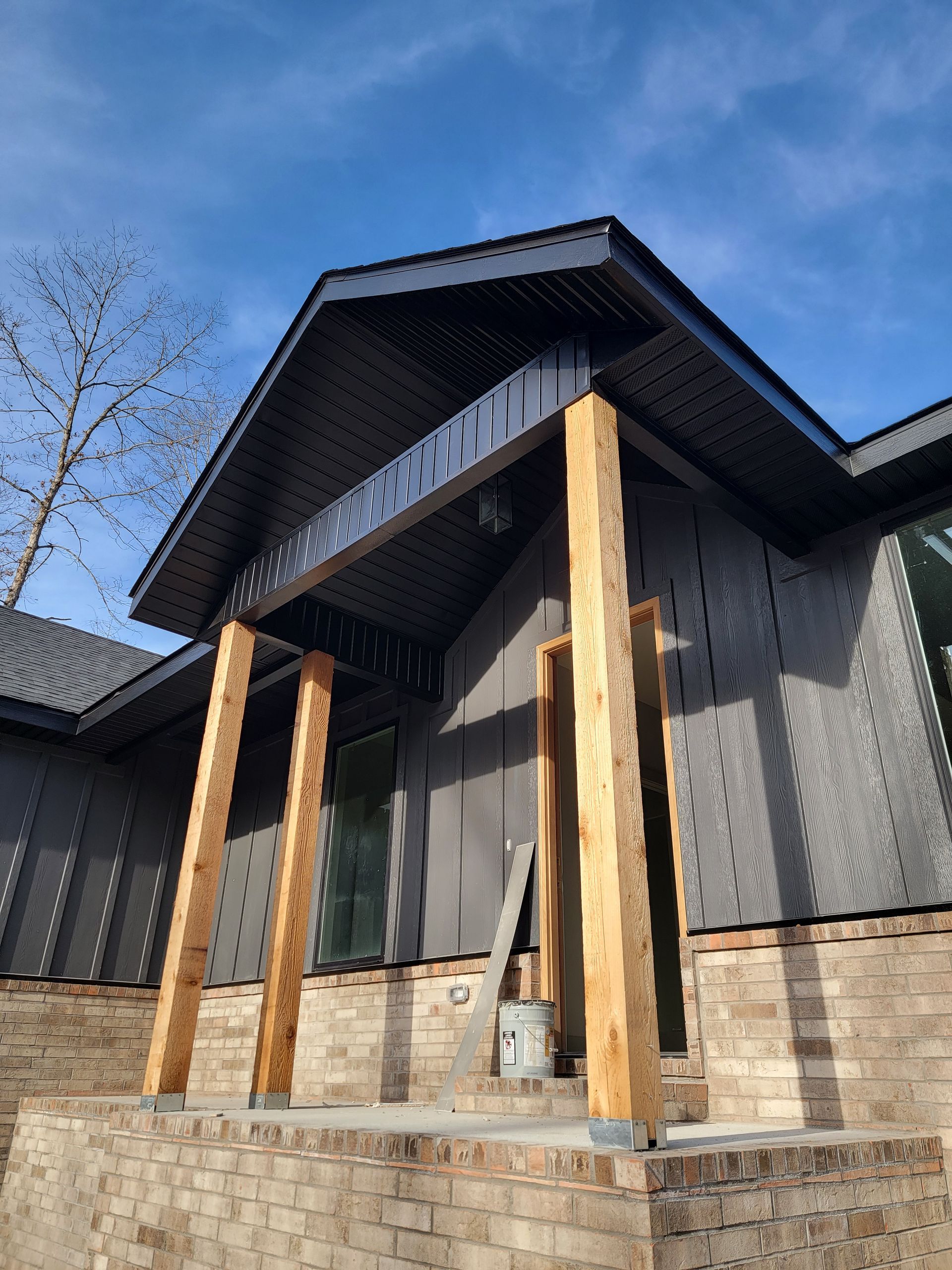 Modern house entrance with stone steps, tall wooden posts, and dark siding under a blue sky