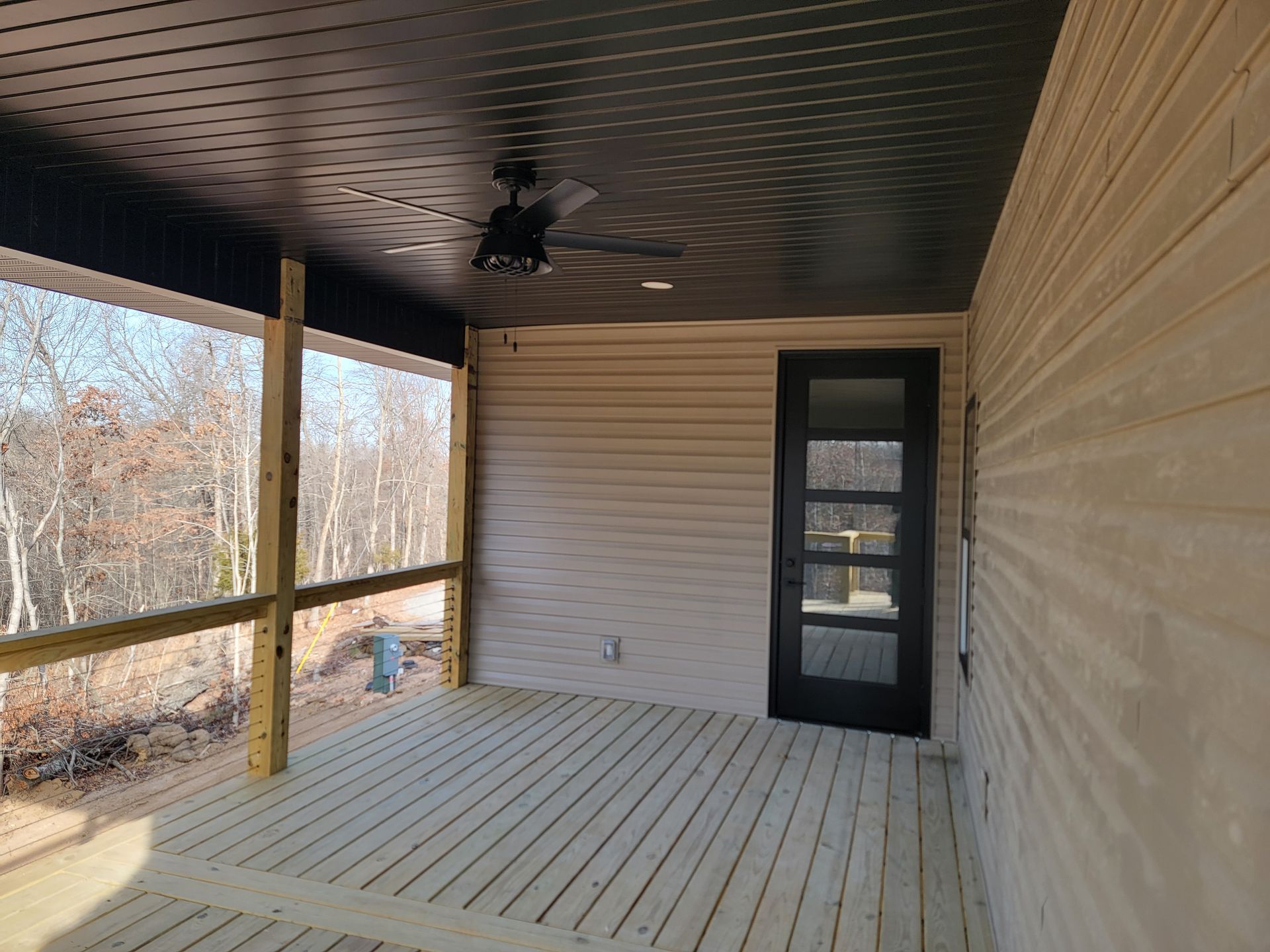 Covered porch with wood siding, ceiling fan, and black door overlooking a wooded yard