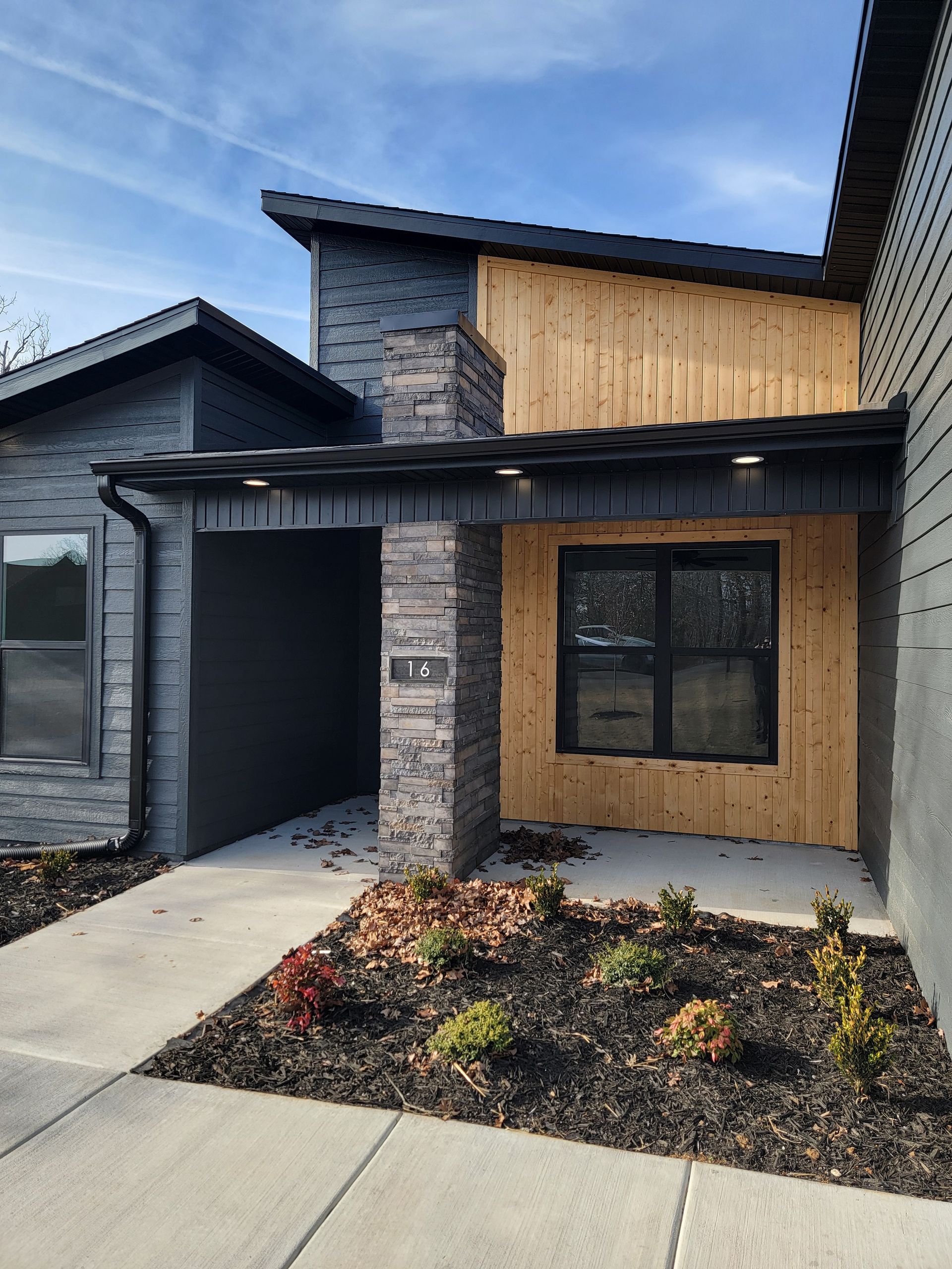 Modern house exterior with stone and wood facade, covered entry, and landscaped front bed.