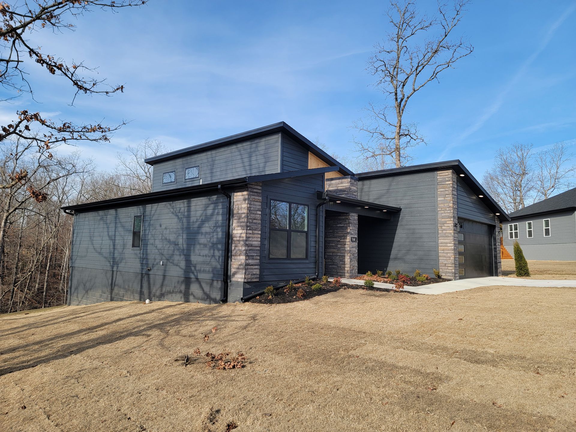 Modern gray two-story house with a sloped driveway and sparse trees under a clear blue sky