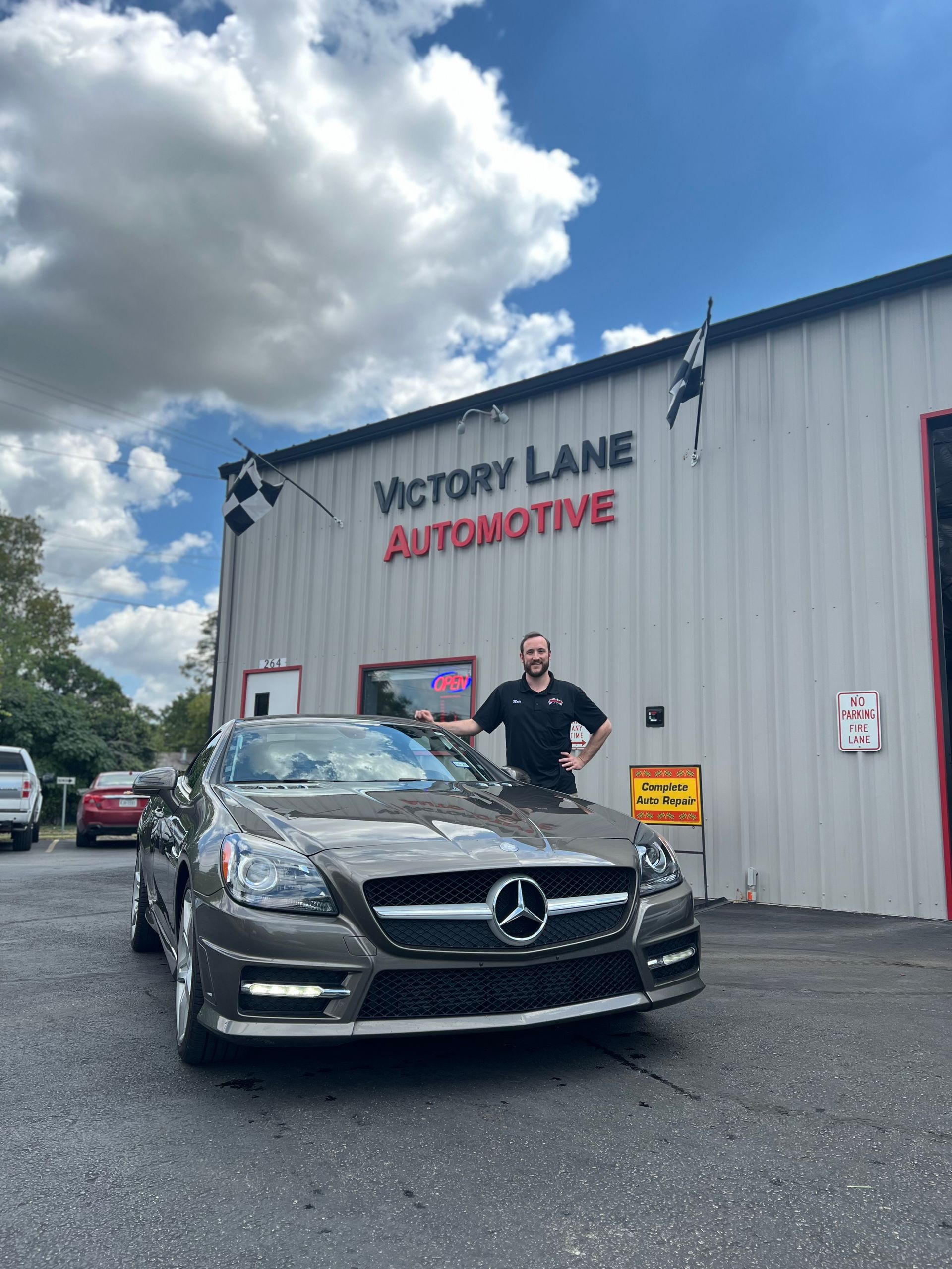 Man poses with gray Mercedes in front of Victory Lane Automotive building. Blue sky. | Victory Lane Automotive