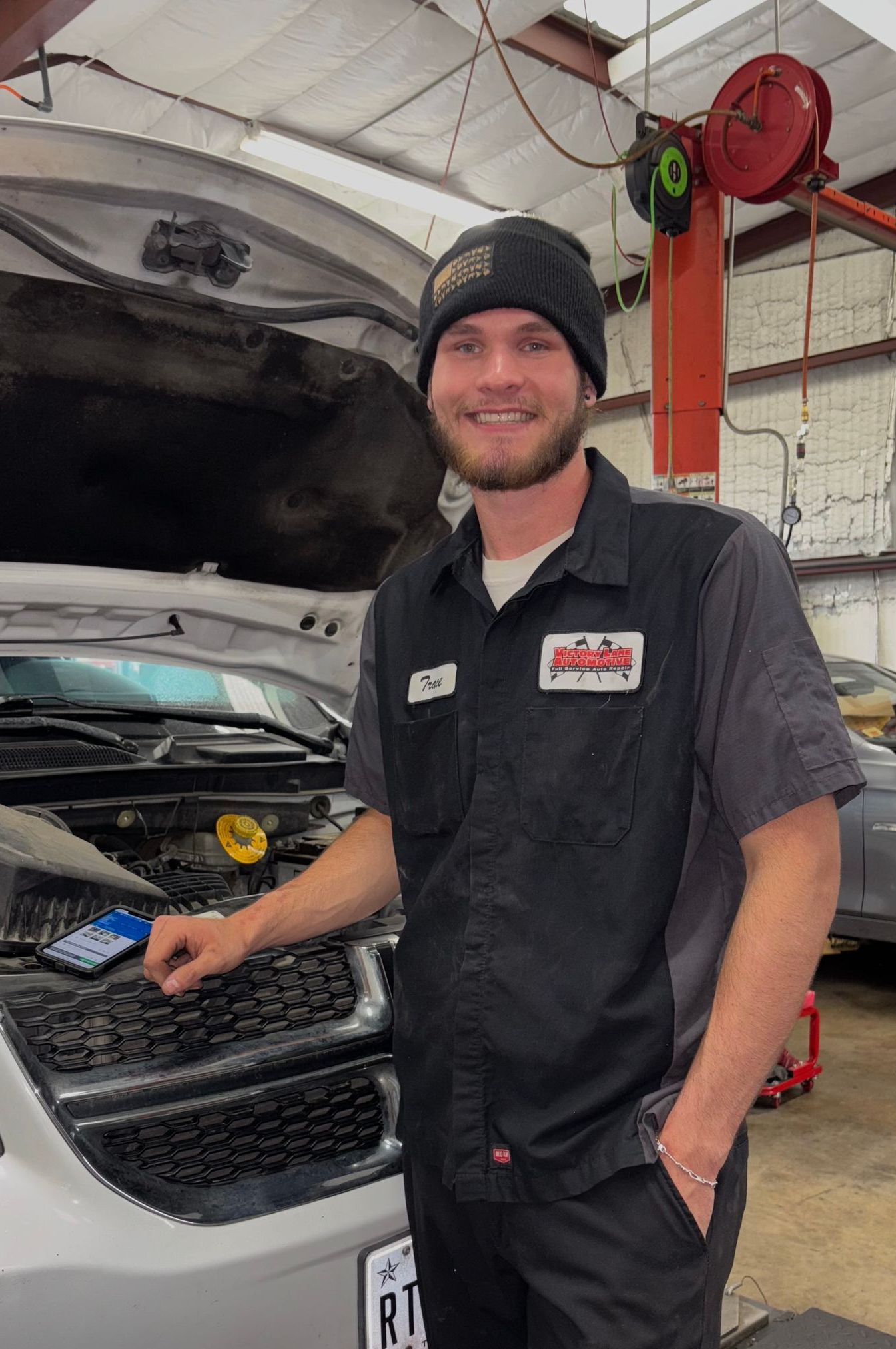 Mechanic in a garage, smiling, working on a car with the hood open. Wearing a work shirt and beanie. | Victory Lane Automotive