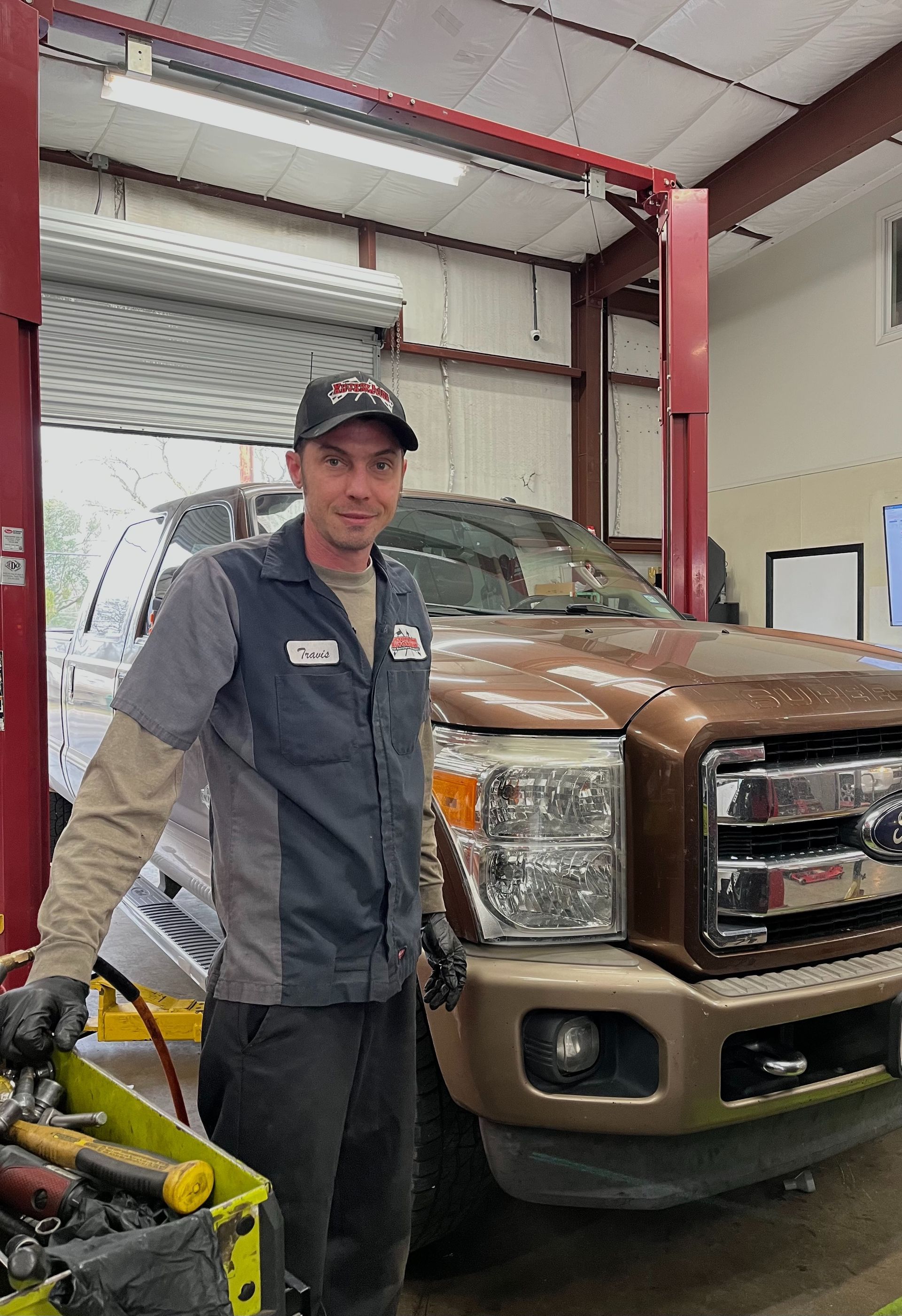Mechanic in uniform stands next to a brown truck in a garage; a toolbox is in the foreground. | Victory Lane Automotive