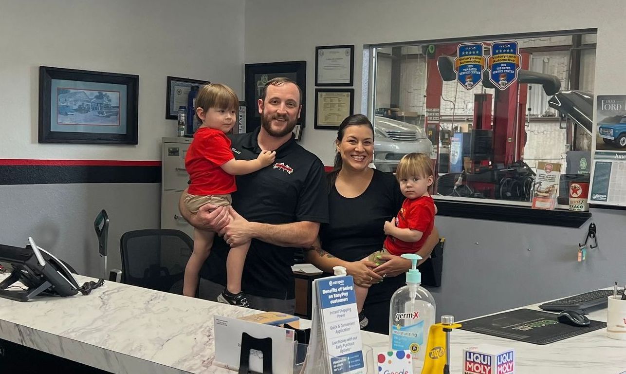Family at auto shop counter, man holding child, woman holding child, smiling, shop interior. | Victory Lane Automotive