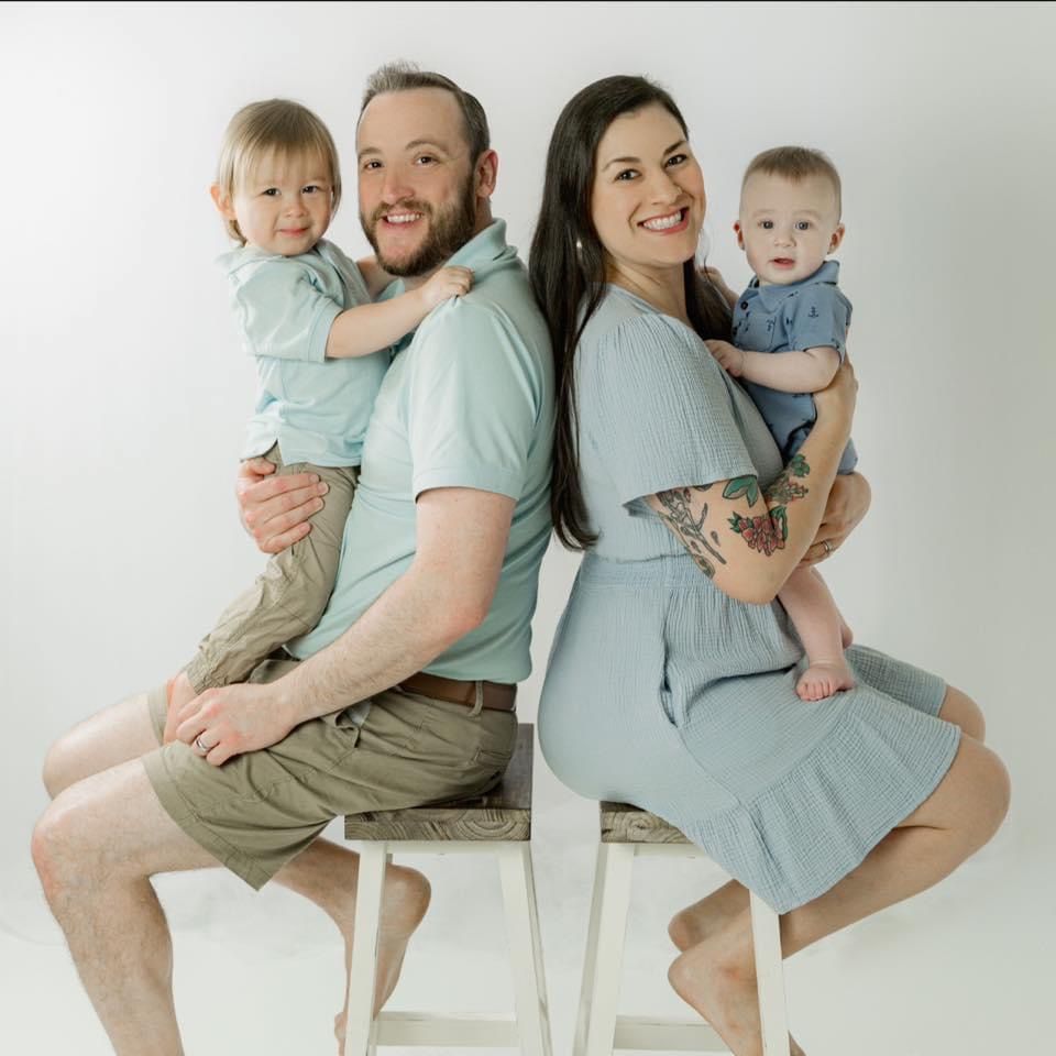 Family of four seated back-to-back. Two parents hold toddlers. Light blue clothing, white background. | Victory Lane Automotive