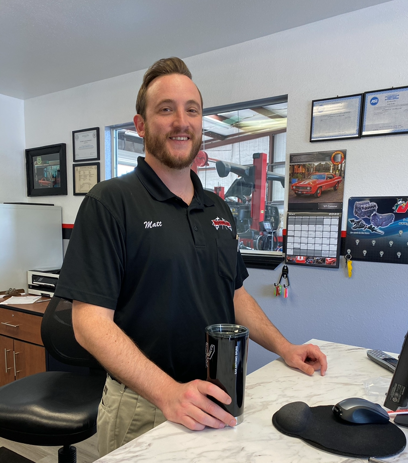 Man in black shirt at a desk, smiling. Shop in background. | Victory Lane Automotive