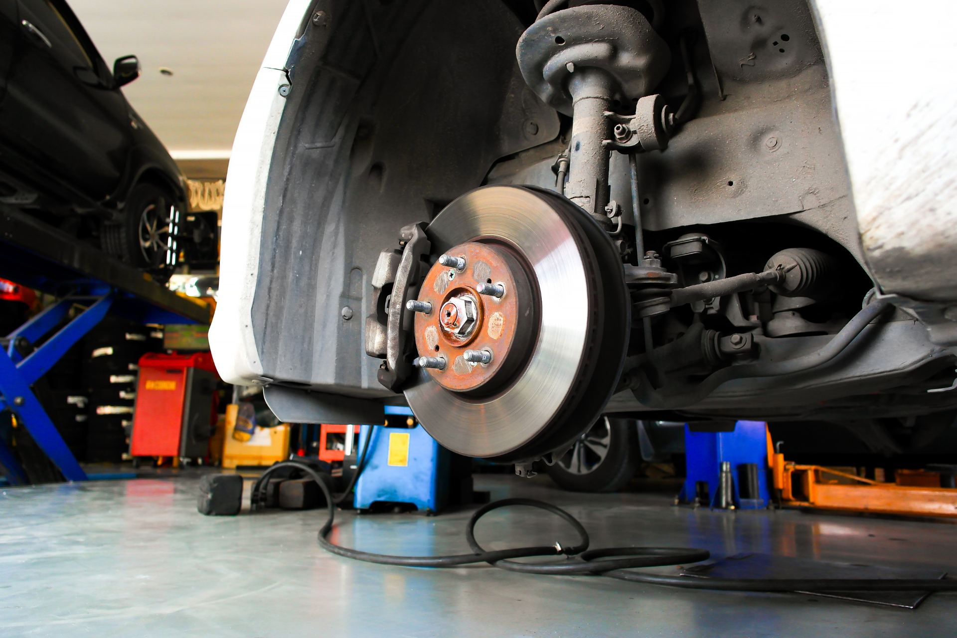 Car's front wheel in a repair shop. Brake rotor and caliper exposed, car on jack, blue lift in background.