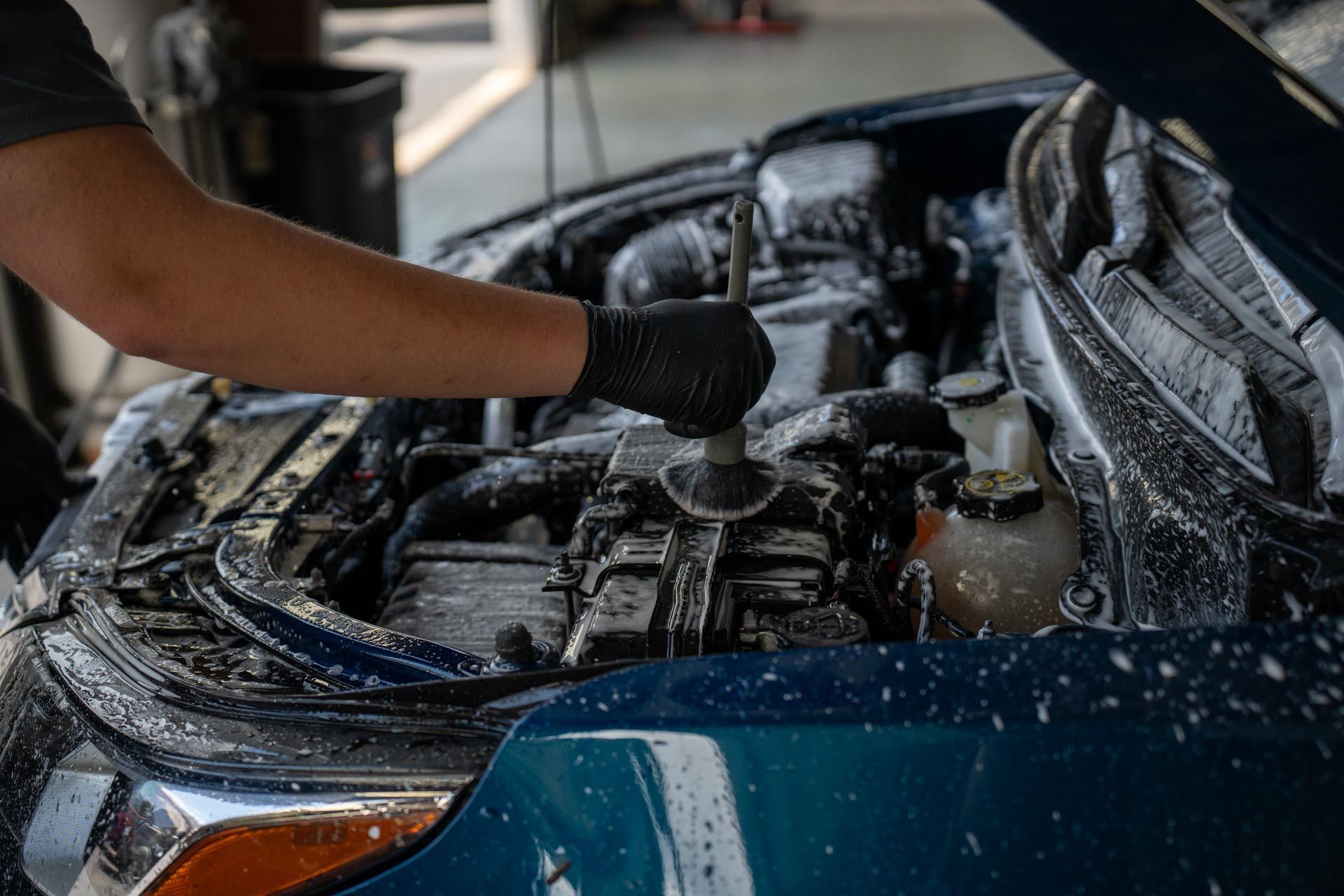 Person cleaning a car engine with soap; blue car, black gloves, garage setting.