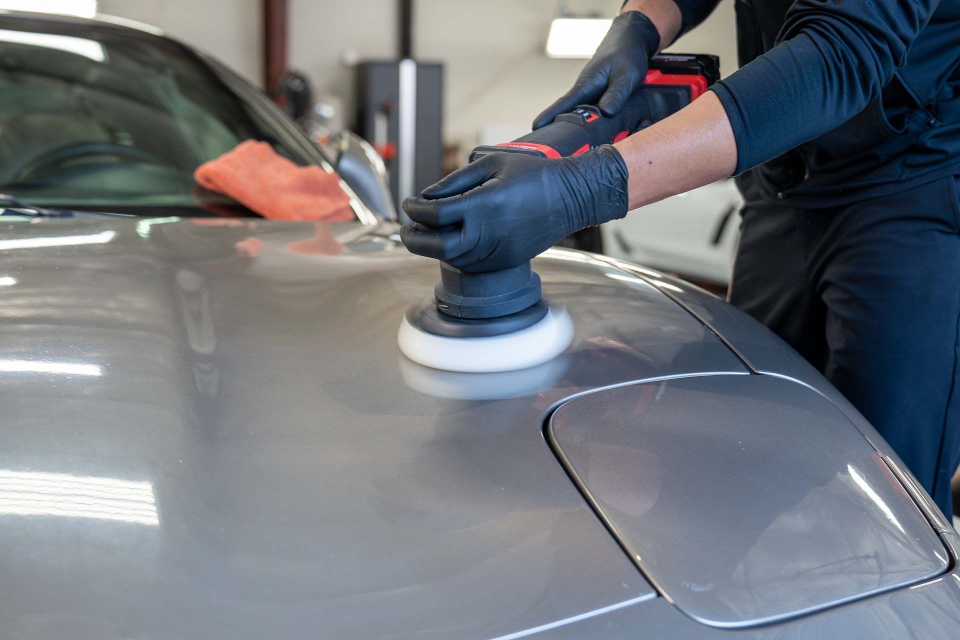 Person in black gloves polishing a gray car hood with a power buffer in a garage.