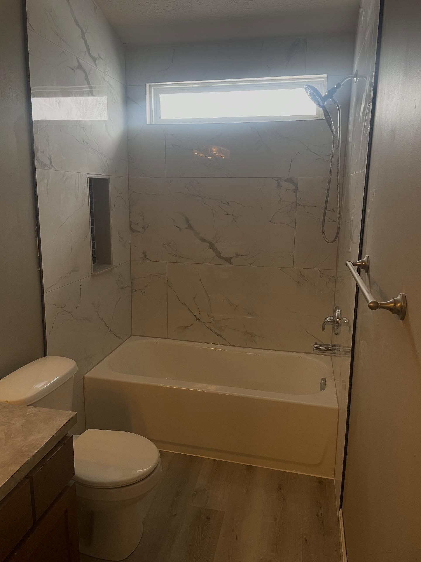 Small bathroom with a bathtub, toilet, marble-patterned walls, and a frosted window above the tub.