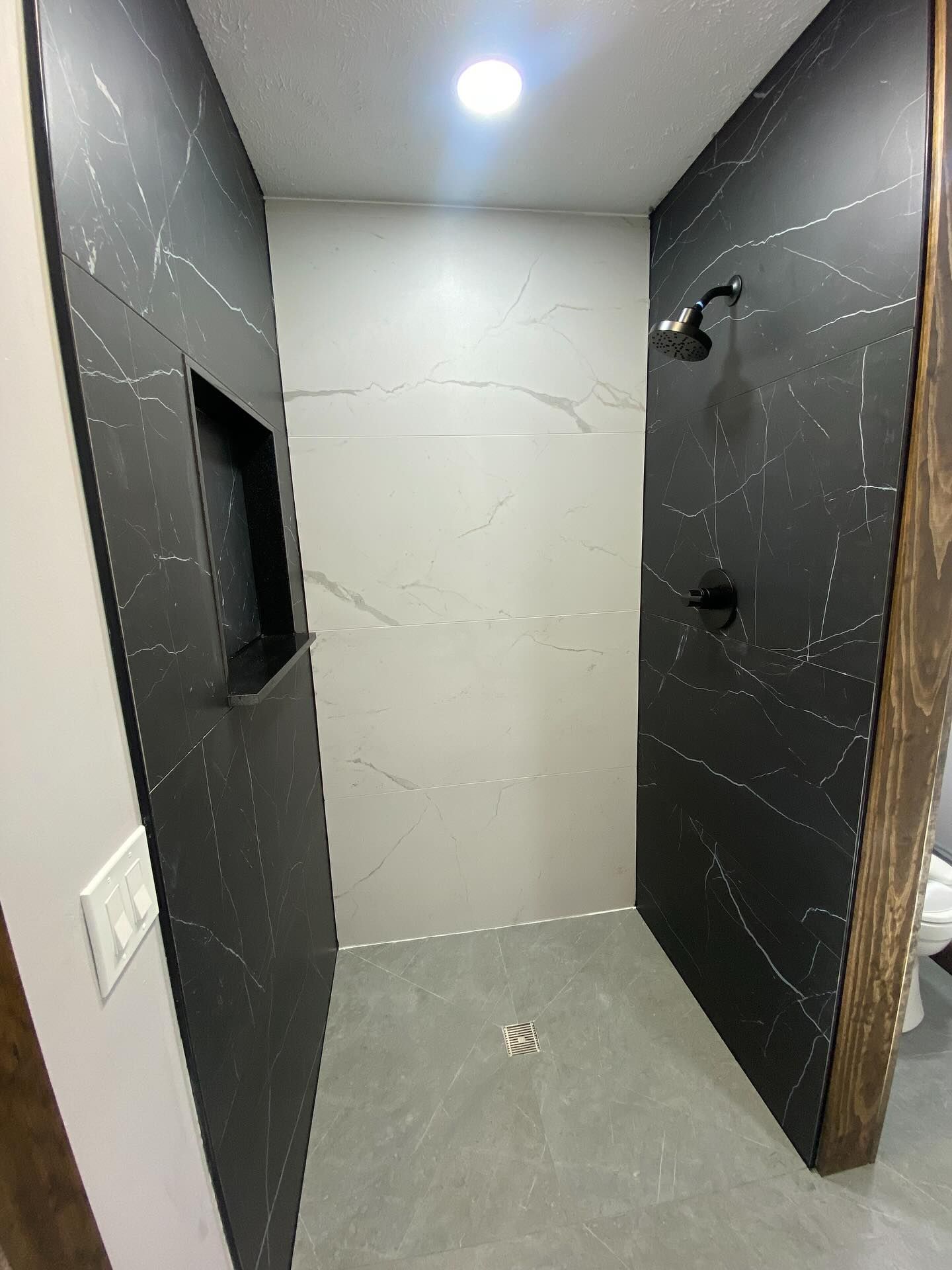 Small tiled shower stall with dark wall panels, a white back wall, and a ceiling light.