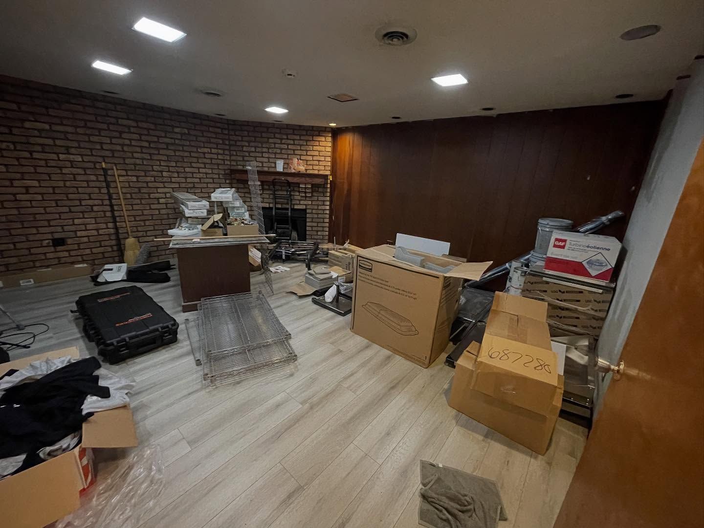Cluttered office or storage room with boxes, chairs, and scattered items on a wood floor