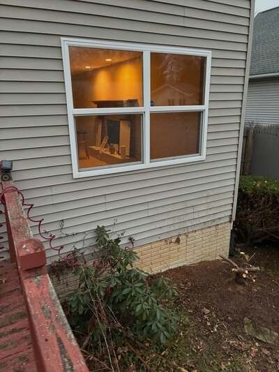 Exterior house wall with a four-pane window, lit warm interior visible inside, and garden bed below.