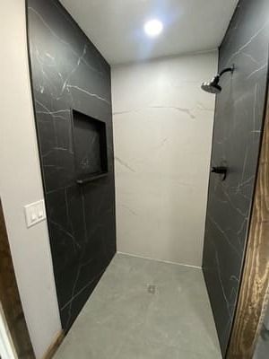 Narrow modern shower stall with dark tile walls, light floor, and recessed shelf niche.
