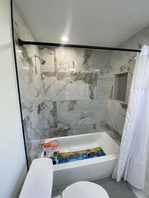Modern bathroom with a marble-tile shower, white tub, and colorful toys inside the tub