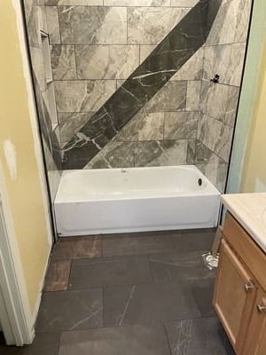 Bathroom with a white tub, gray tile walls, and dark tile floor next to a vanity cabinet.
