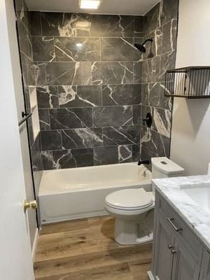 Small bathroom with dark marble shower wall, white tub, toilet, gray vanity, and wood-look floor.