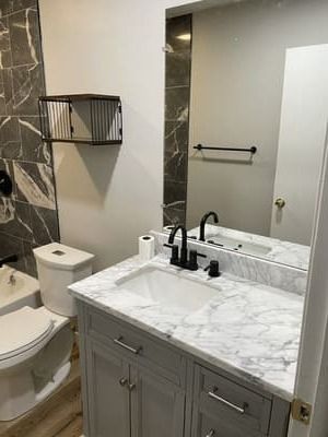 Small bathroom with marble vanity, black faucet, toilet, mirror, towel rack, and wall shelf.