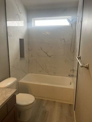 Small bathroom with a bathtub, toilet, vanity, and window above marble-look walls.
