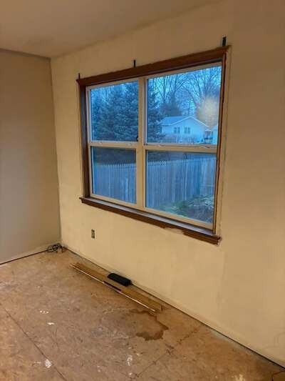 Empty room with beige walls, wooden-framed window, and bare floor under renovation.