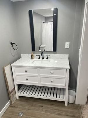 White bathroom vanity with black mirror, faucet, and slatted shelf in a gray-walled room.