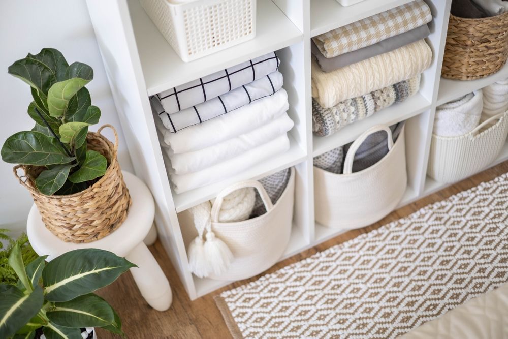 Neatly organized white shelving with rolled towels, baskets, and a green potted plant beside a patterned rug