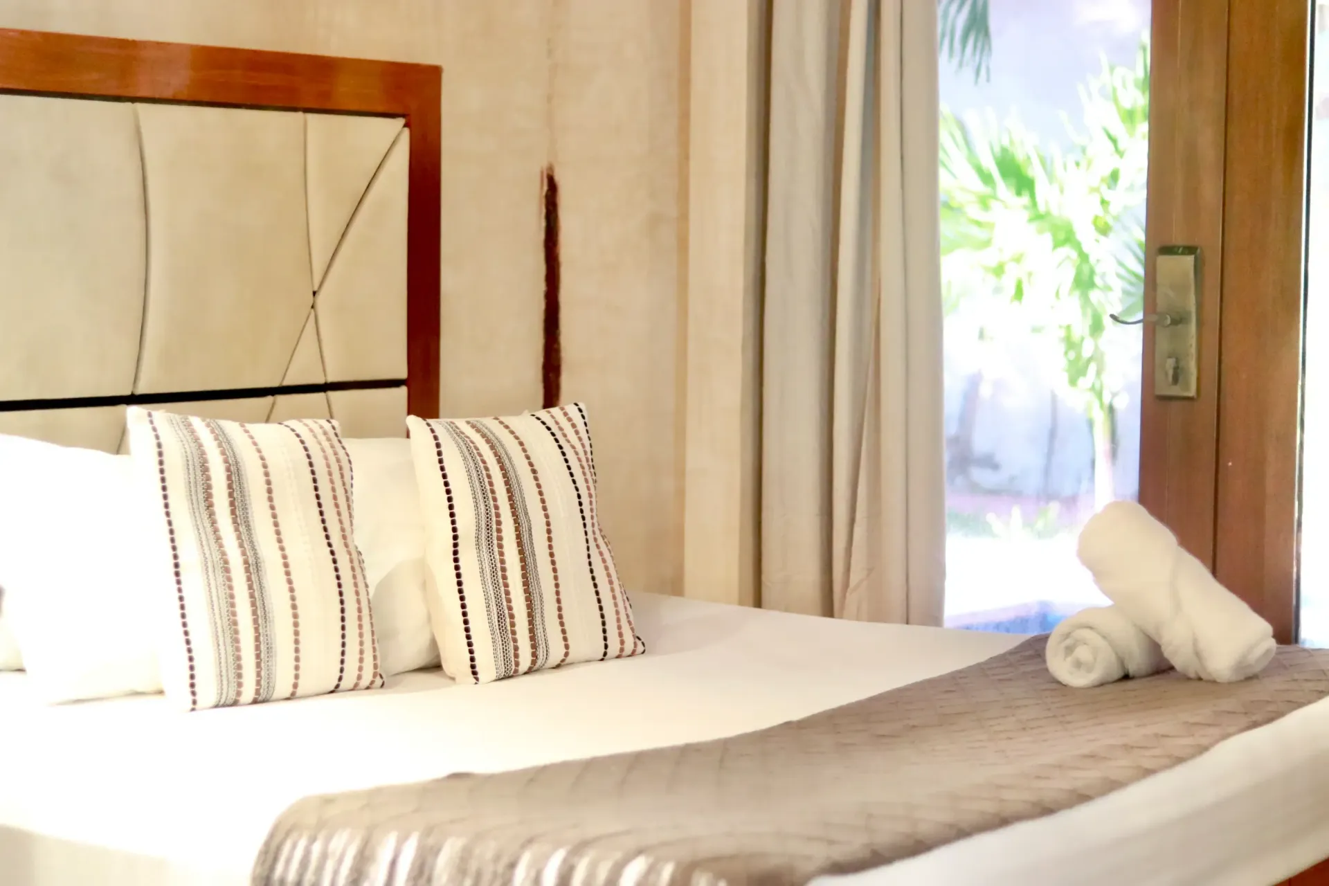Neatly made bed with striped pillows in a bright, sunlit bedroom near a window and curtains
