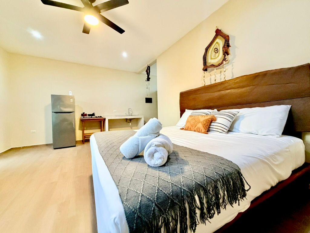 Bright bedroom with a wooden bed, gray throw, blue towels, desk, and mini fridge under ceiling fan